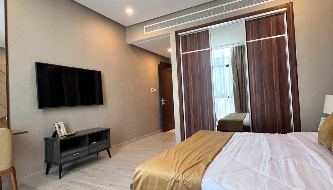 Modern bedroom with a wooden bed, matching wardrobe, wall-mounted TV, and light-colored flooring; neutral walls and minimal decor.