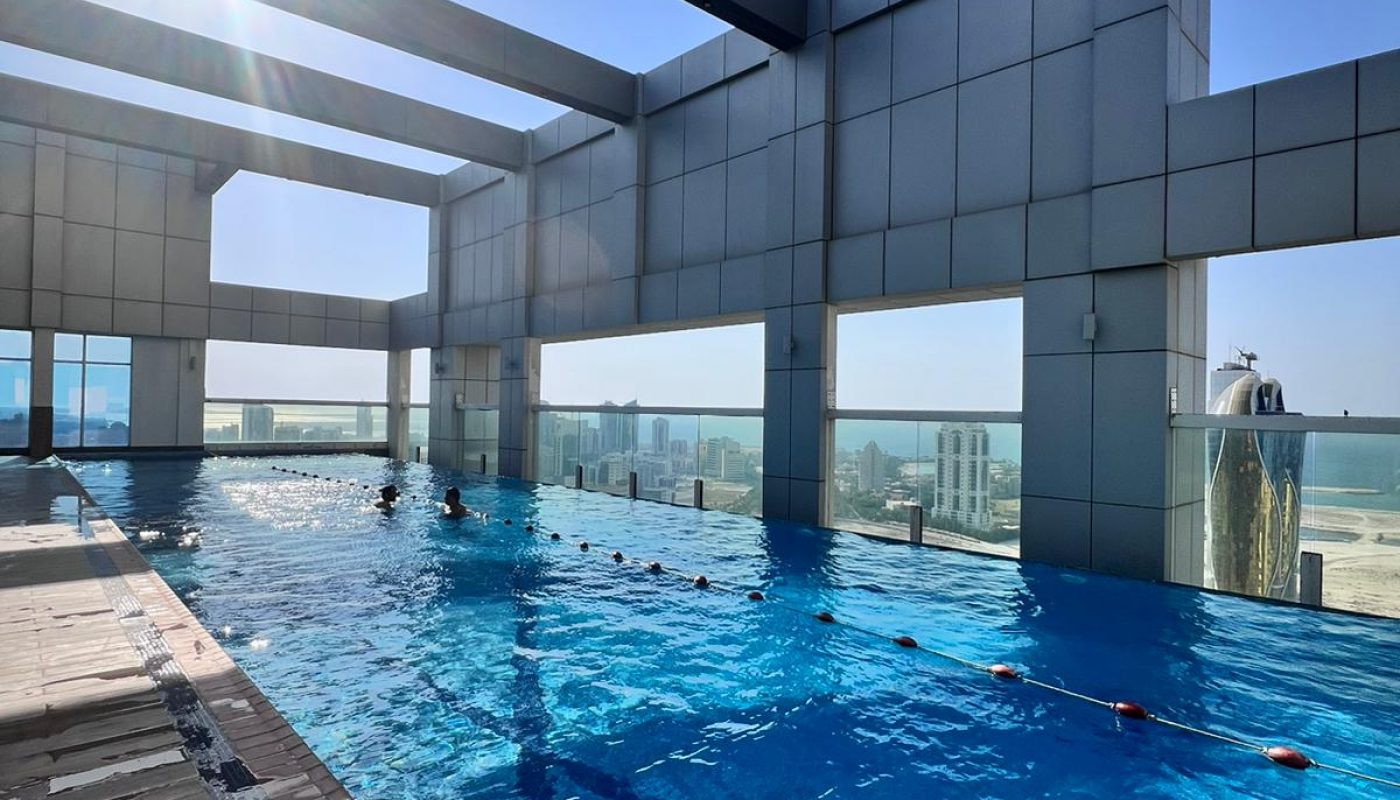 Indoor rooftop swimming pool with clear blue water, sun shining through open beams, and a cityscape visible in the background.