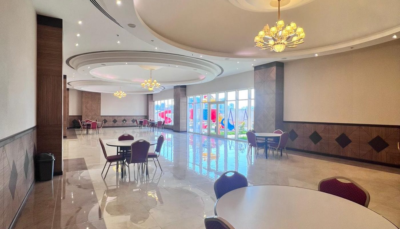 A spacious, modern room with round tables and red chairs, polished marble floors, large ceiling lights, and colorful windows along one side.