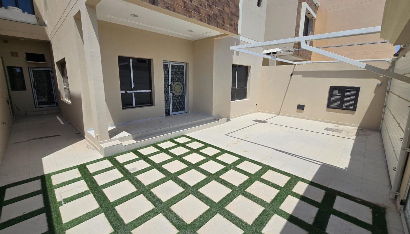 A modern house with a beige exterior features a tiled patio with grass strips, windows, and a decorative front door. Located in Damistan, this new villa offers a yard with a mix of pavement and grassy patches and is currently for sale.