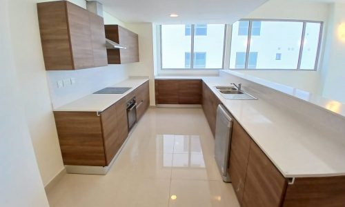 A modern kitchen in a flat for rent in the Amwaj area, featuring wooden cabinets, a built-in oven and stove, a sink, a dishwasher, and large windows providing natural light. The floor is tiled, and the countertops are white. This rental property offers everything you need for comfortable living.