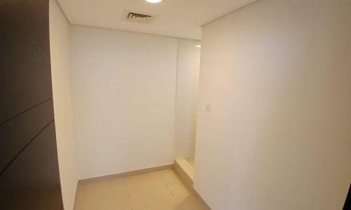 A small, unfurnished flat for rent in the Amwaj area features a white-walled room with beige tiled flooring, a white ceiling, a recessed light, and a closed door partially visible on the left side.