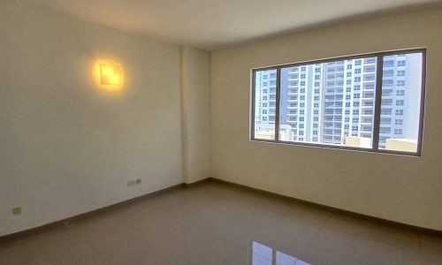 A bare room with a tiled floor, a lit wall light, and a large window showcasing a view of tall buildings outside. This flat for rent in the Amwaj area offers modern living with urban scenery.