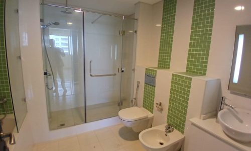 A modern bathroom in this Amwaj rental property features a glass-enclosed shower, a wall-mounted toilet and bidet, a sink with a mirror, and green tiled accents on the walls. Perfect for those seeking sophisticated living in a flat for rent.