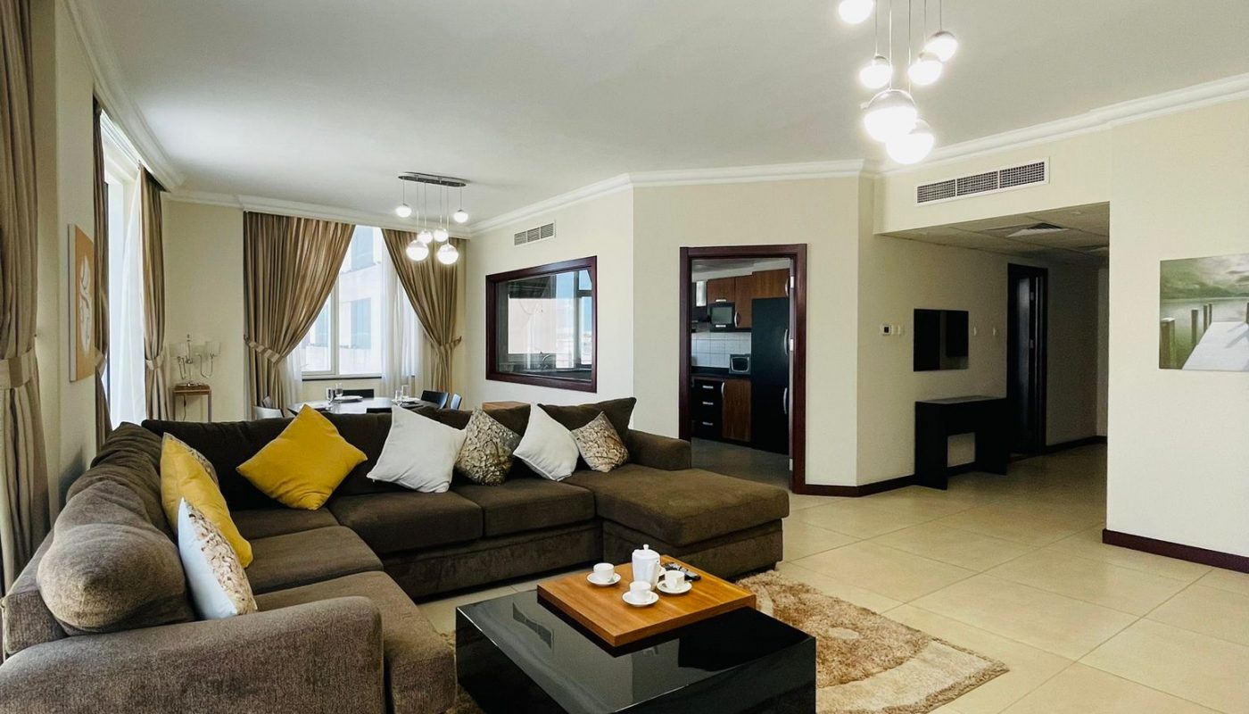 Spacious living room with a sectional sofa, coffee table, rug, neutral decor, large windows, and modern lighting. Open view to kitchen and hallway.