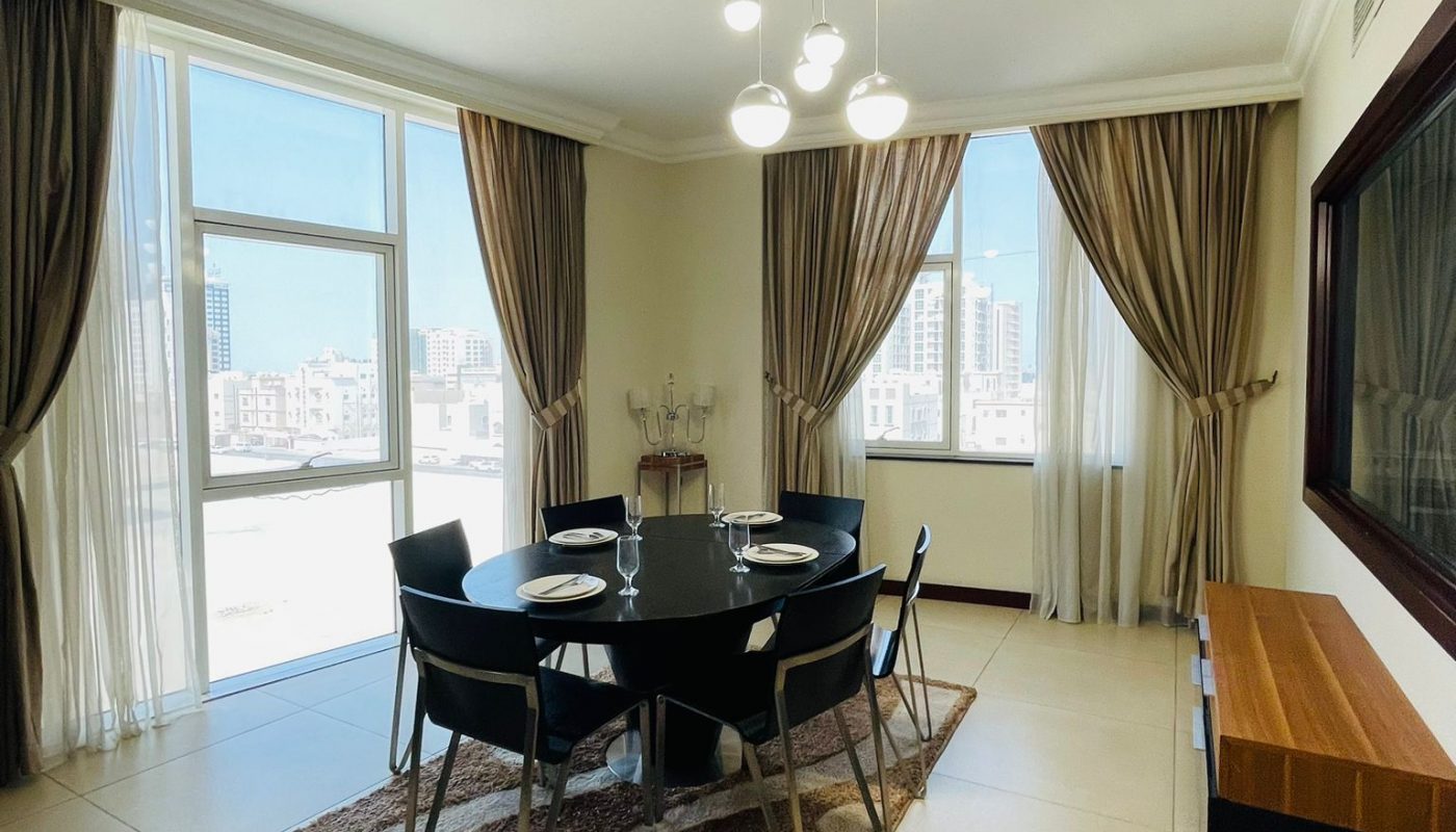A modern dining room with a round black table, six chairs, large windows with curtains, a rug, a sideboard, and a wall mirror, with city buildings visible outside.