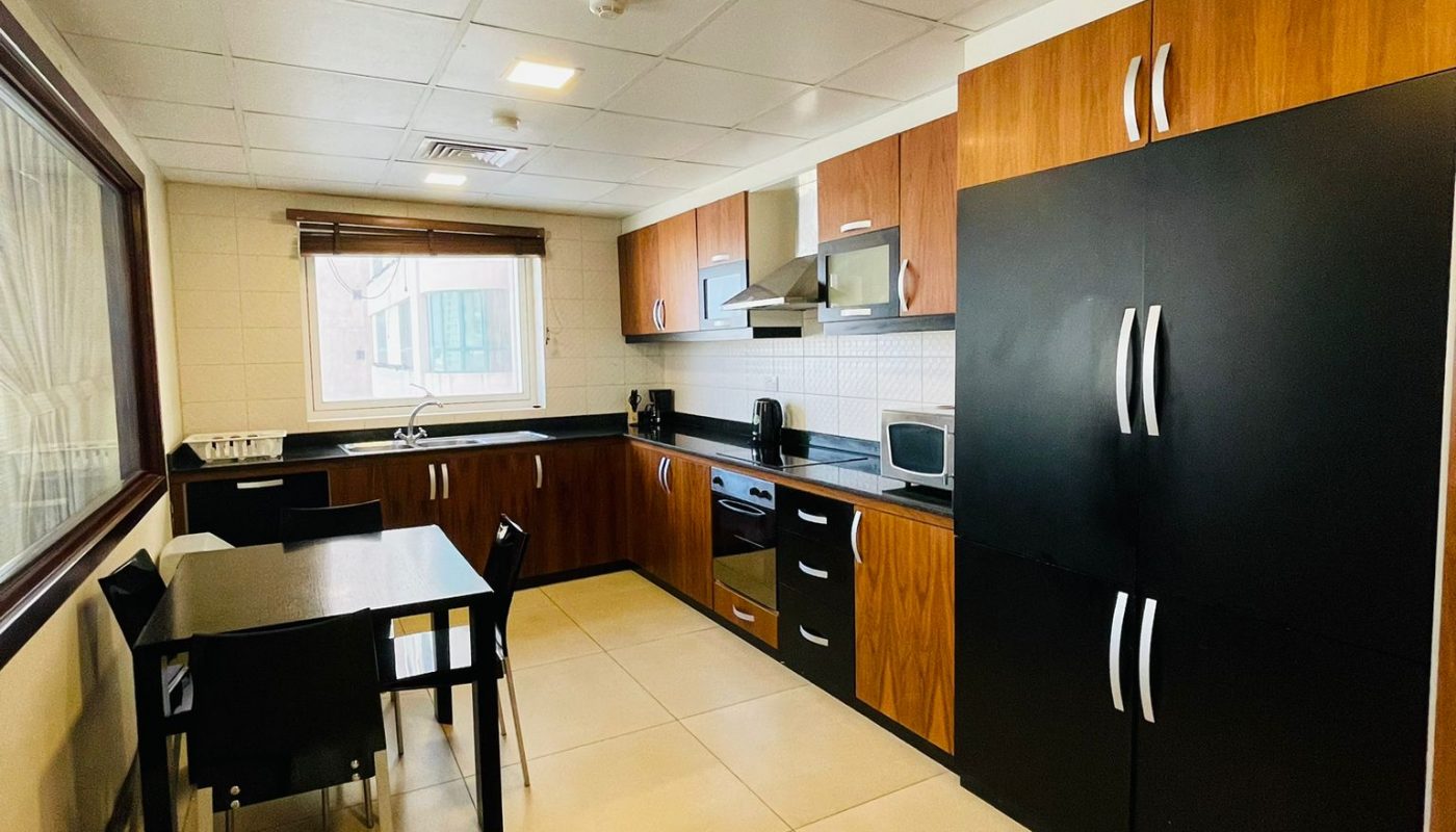Modern kitchen with wood and black cabinets, a dining table with four chairs, tiled floor, and appliances including a stove, microwave, and refrigerator. Large window on the left wall.
