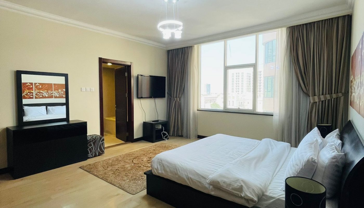 A modern bedroom with a large bed, bedside table, dresser with mirror, mounted TV, window with city view, and light wood flooring.
