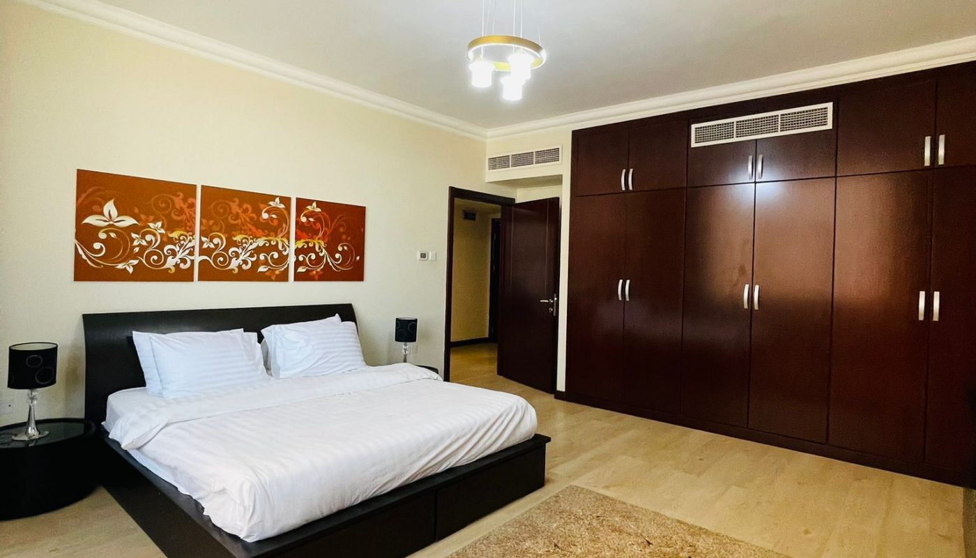 A spacious bedroom with a large bed, white bedding, a beige rug, wooden wardrobes, and modern wall art above the bed.
