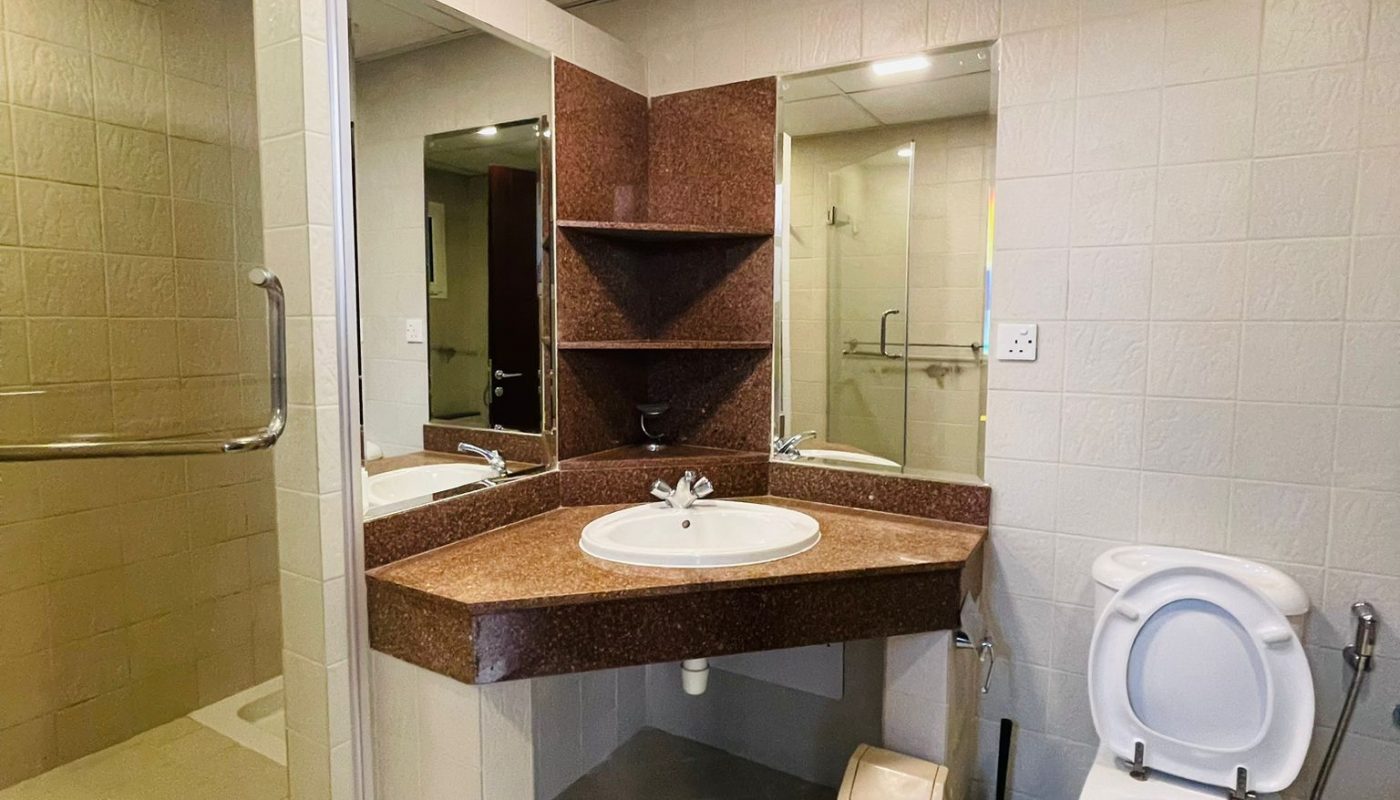 A bathroom with a glass-enclosed shower, a corner sink with shelves, a mirror, a toilet, a trash bin, and a toilet brush on a tiled floor.