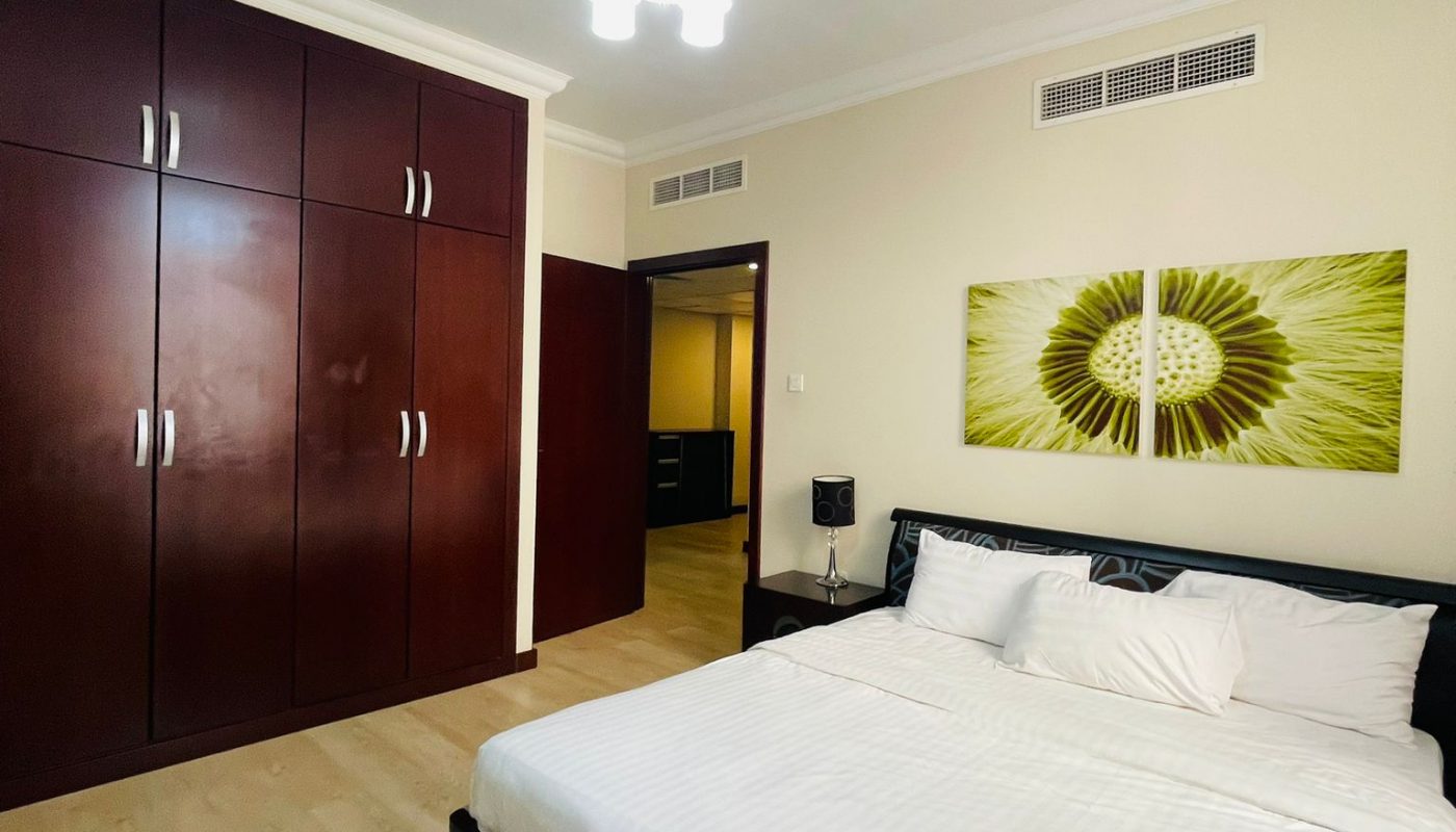 A modern bedroom with a double bed, white bedding, a dark wooden wardrobe, light wood flooring, and a green abstract artwork above the bed.