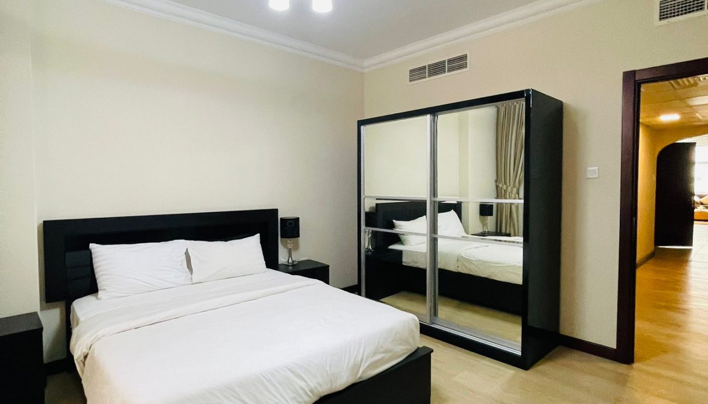 A modern bedroom with a double bed, white bedding, two bedside tables, a mirrored wardrobe, and light wood flooring under a ceiling light fixture.