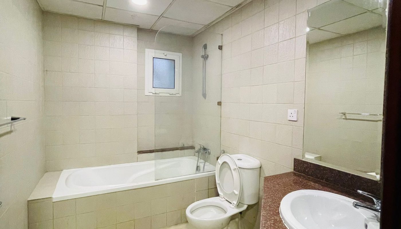 A bathroom with beige tiles, a bathtub with a glass partition, a toilet, a wall-mounted sink with a brown marble countertop, a mirror, and a ceiling light.