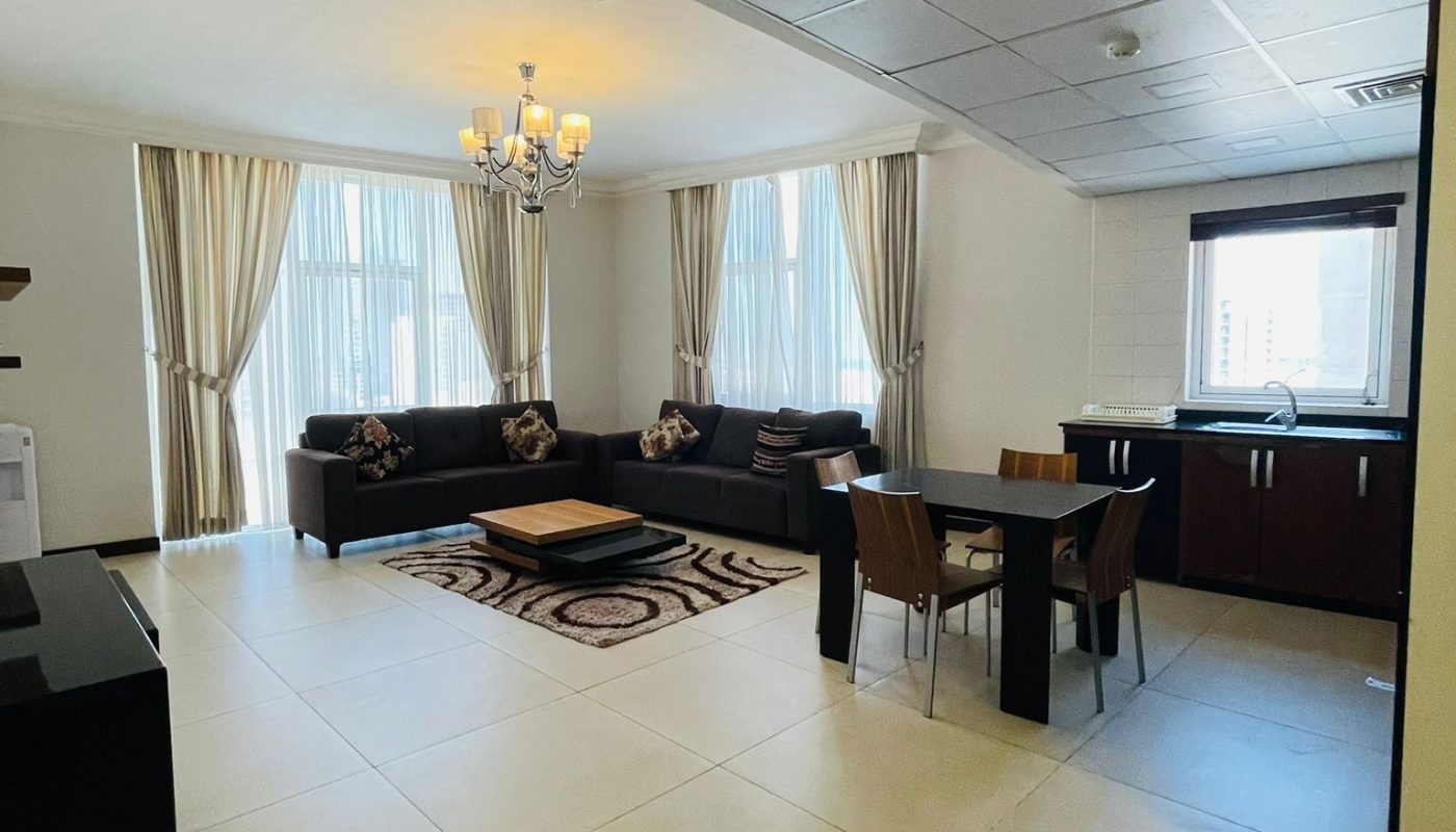 Spacious living and dining area with tiled floors, large windows with curtains, a black sofa set, a rug, dining table with chairs, and an open kitchen.