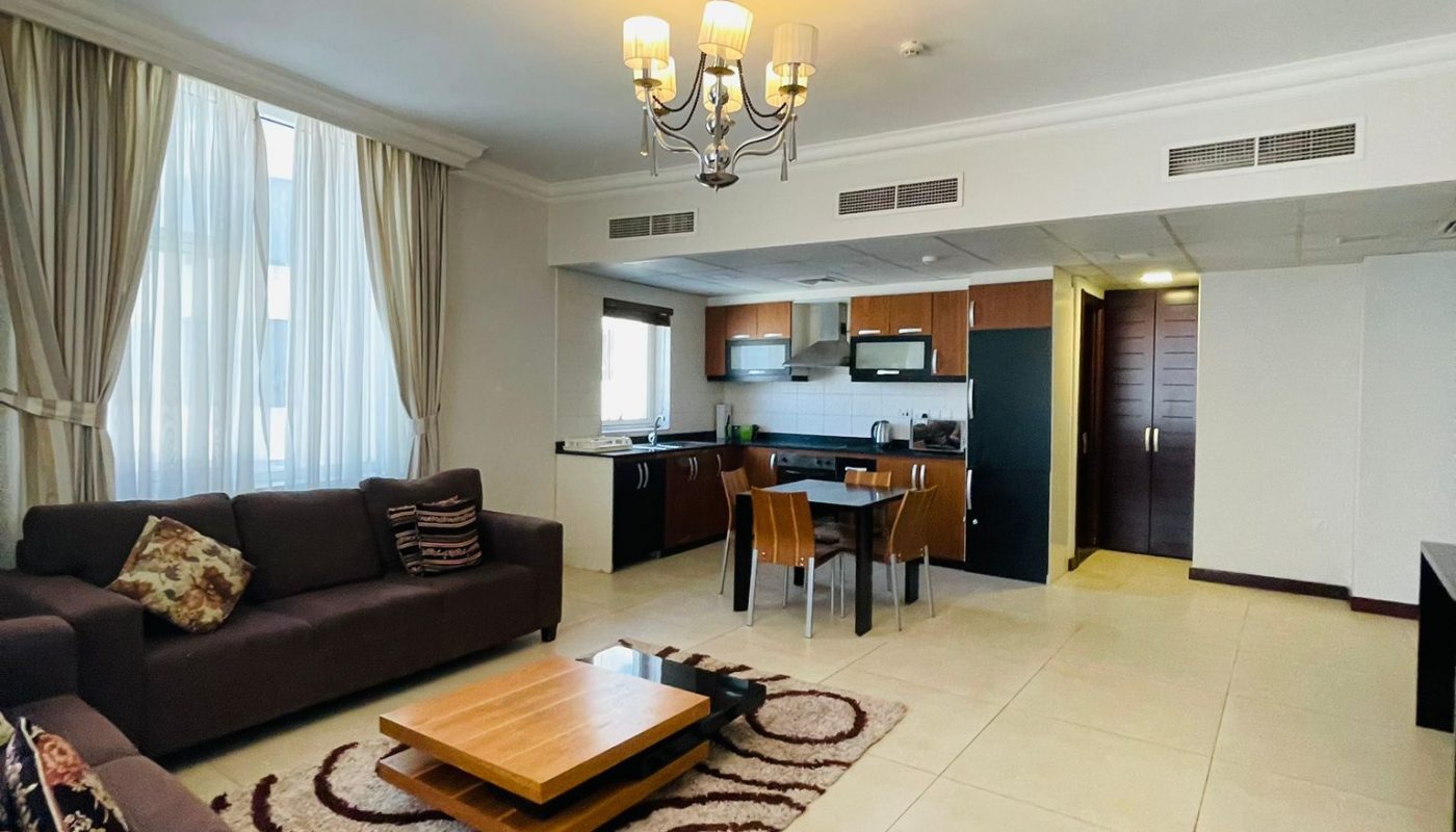Open-plan living and dining area with dark sofas, a wooden coffee table, patterned rug, and a kitchen in the background under modern ceiling lights.