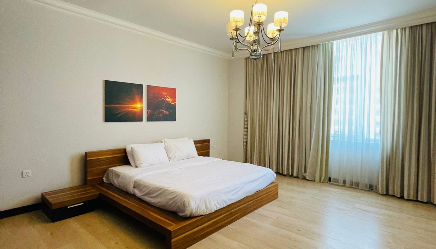 A spacious bedroom with a wooden bed, white bedding, two sunset artworks above the bed, a chandelier, large window with curtains, and light wood flooring.