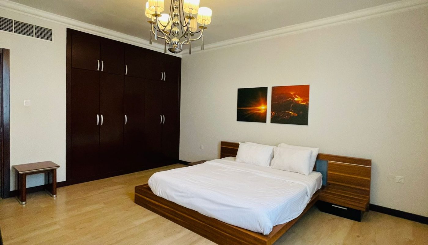 A minimalist bedroom with a wooden bed, white bedding, two nightstands, a wall-mounted wardrobe, light wood flooring, and a ceiling chandelier. Two pictures hang above the bed.