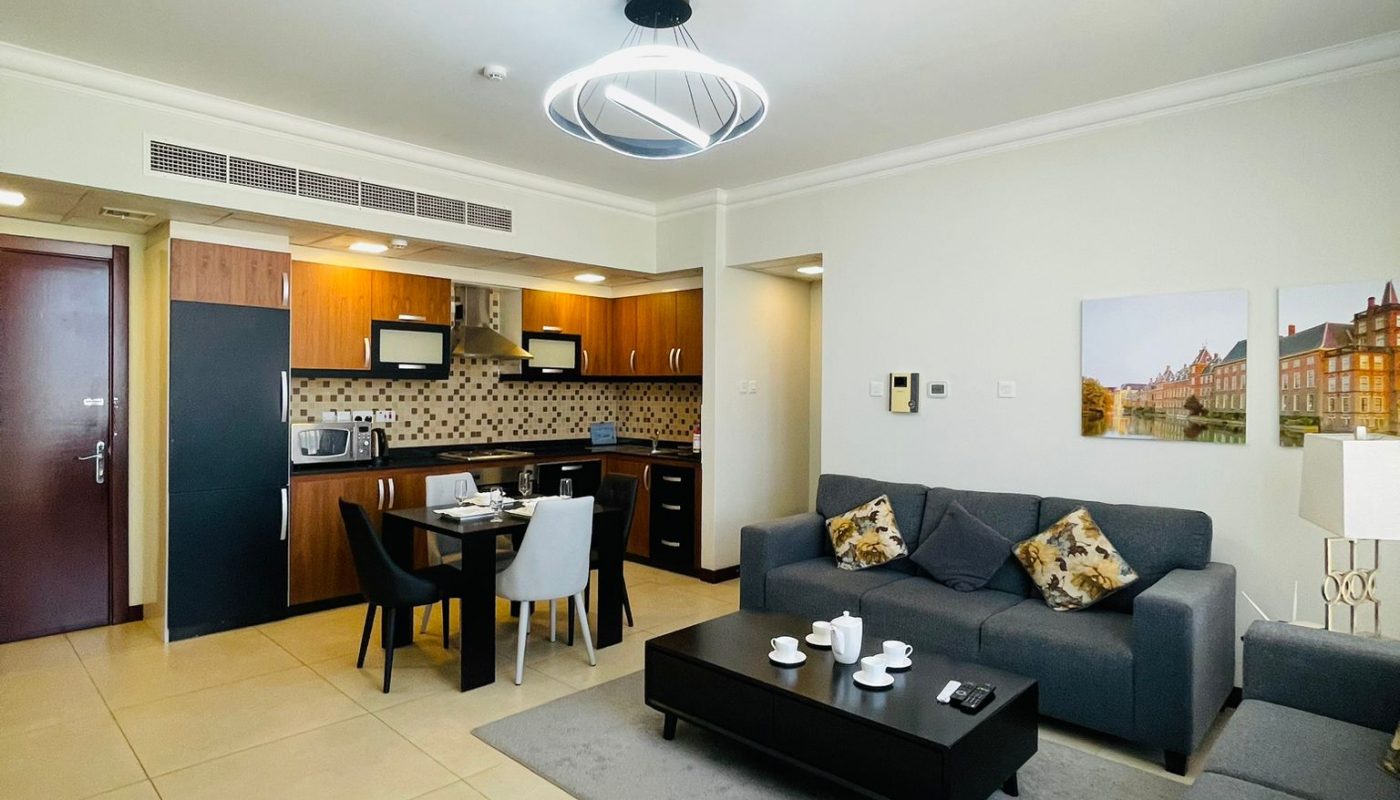 Modern open-plan living room and kitchen area with a gray sofa, coffee table, dining set, and wooden cabinets under bright lighting.