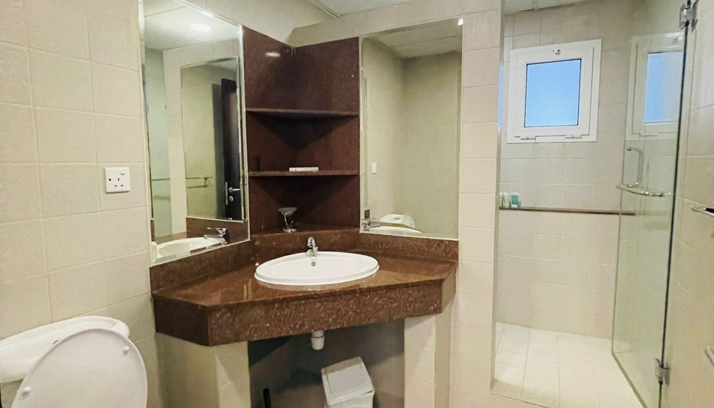 Bathroom with a white toilet, sink set in a brown countertop, mirror, shelves, waste bin, glass shower enclosure, and two small windows.