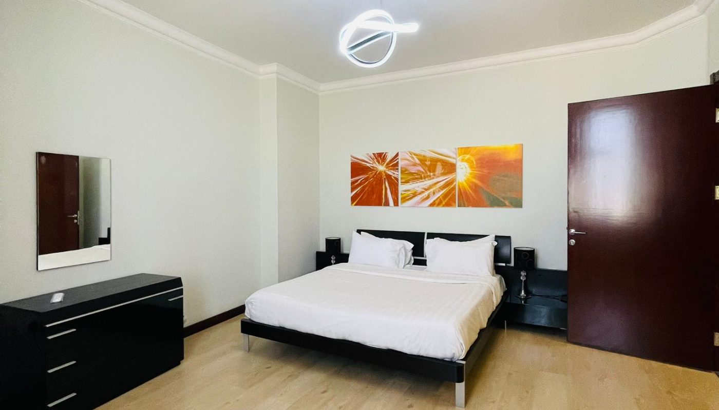 Minimalist bedroom with a double bed, two white pillows, black nightstands, a dresser with mirror, wooden floor, and two orange abstract paintings above the bed.