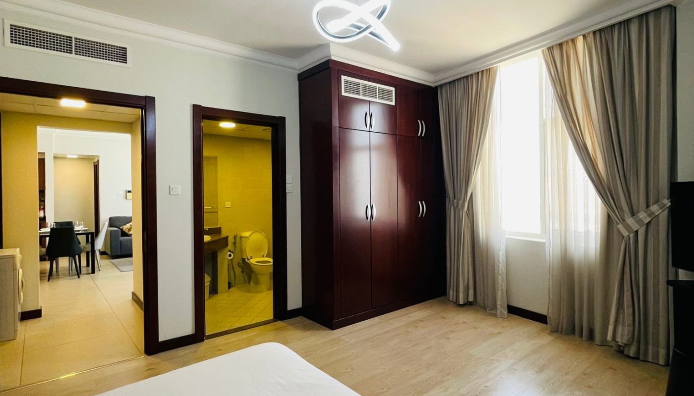 A spacious bedroom with a bed, wooden wardrobe, large window with curtains, visible bathroom, and hallway leading to a living area.