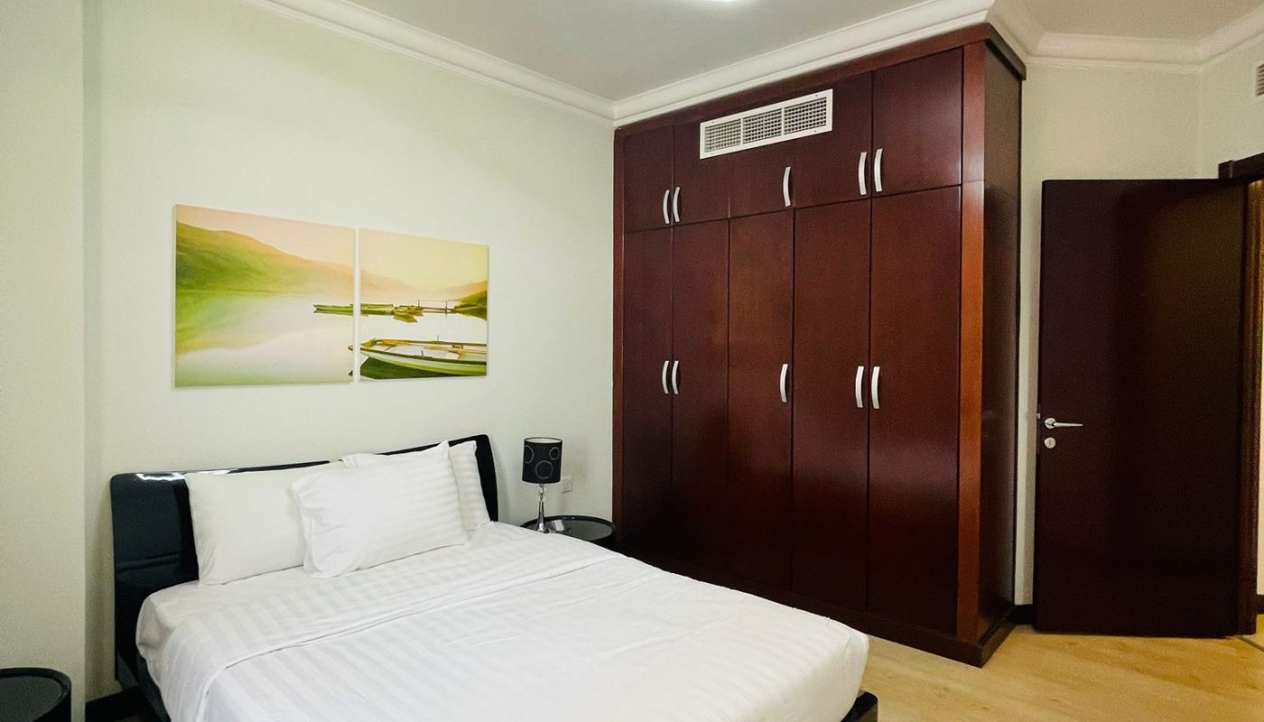 A modern bedroom with a white double bed, wooden floor, dark wood wardrobe, bedside tables, and a ceiling light fixture. A landscape painting hangs above the bed.