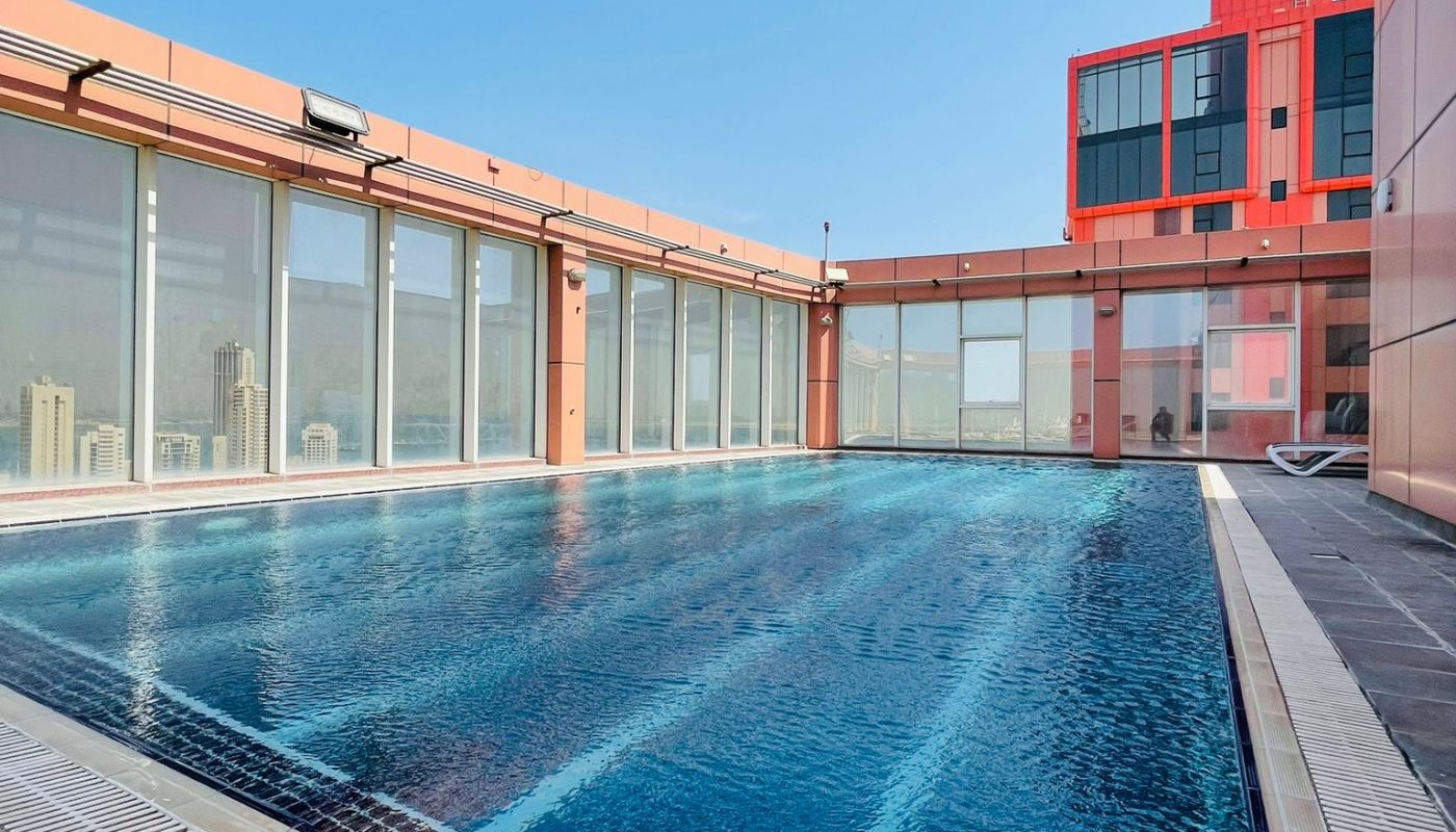 Outdoor rooftop swimming pool with clear blue water, surrounded by a modern building with glass walls and red accents under a clear sky.