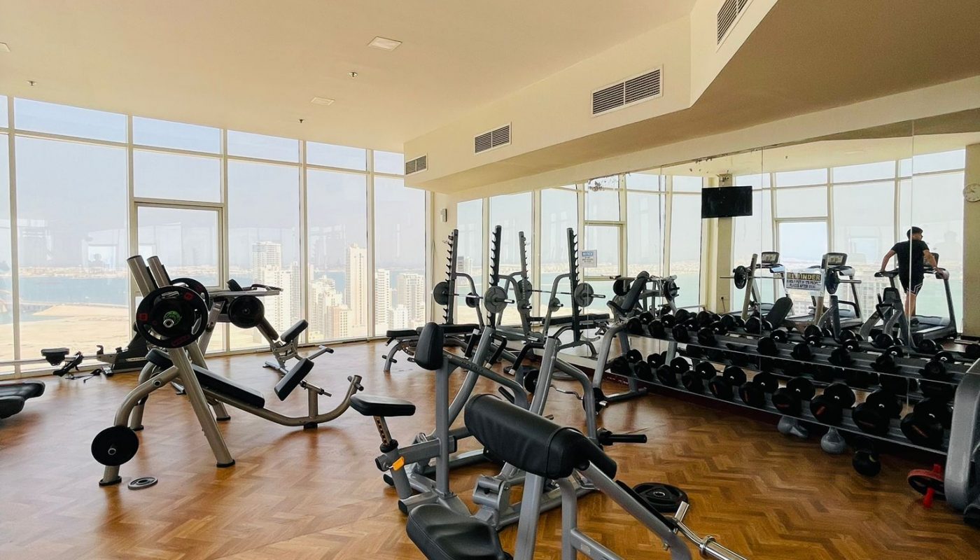 A modern gym with various exercise equipment, mirrors on one wall, large windows with a city view, and wooden parquet flooring.