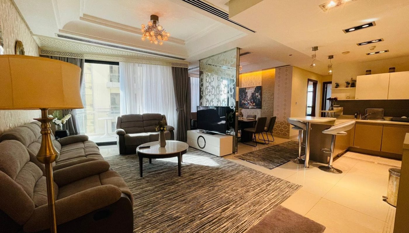 A modern living room with a gray sofa set, coffee table, and a floor lamp in this luxury apartment. The room extends into an open kitchen and dining area with a long counter and bar stools. Large windows bring in natural light, enhancing the serene ambiance of your 2 BR apartment on Reef Island.