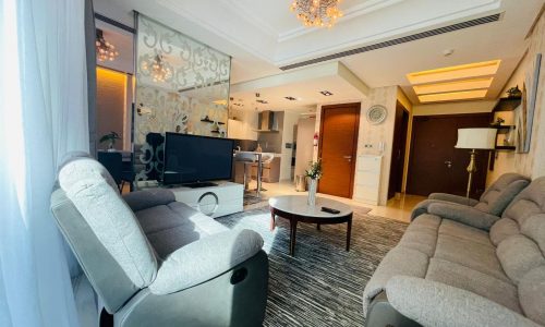 A modern living room in a luxury apartment featuring two gray recliner chairs, a matching sofa, a round coffee table, a TV, an open kitchen in the background, and decorative lighting on the ceiling.