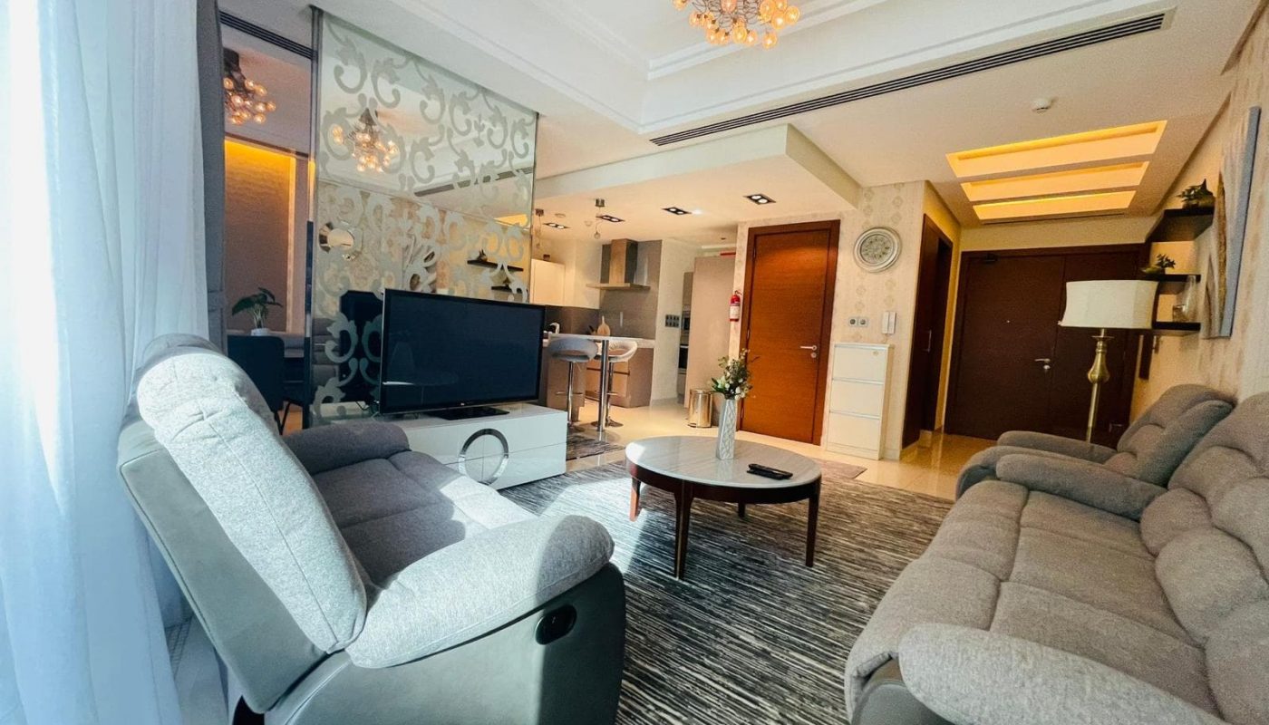 A modern living room in a luxury apartment featuring two gray recliner chairs, a matching sofa, a round coffee table, a TV, an open kitchen in the background, and decorative lighting on the ceiling.