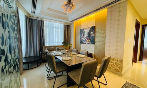 A modern dining room in a luxury apartment on Reef Island features a table set for six, upholstered chairs, a chandelier, and a cityscape painting on the wall. Floor-to-ceiling curtains cover windows near a circular side table.