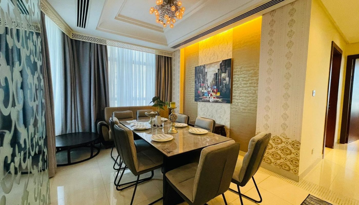 A modern dining room in a luxury apartment on Reef Island features a table set for six, upholstered chairs, a chandelier, and a cityscape painting on the wall. Floor-to-ceiling curtains cover windows near a circular side table.