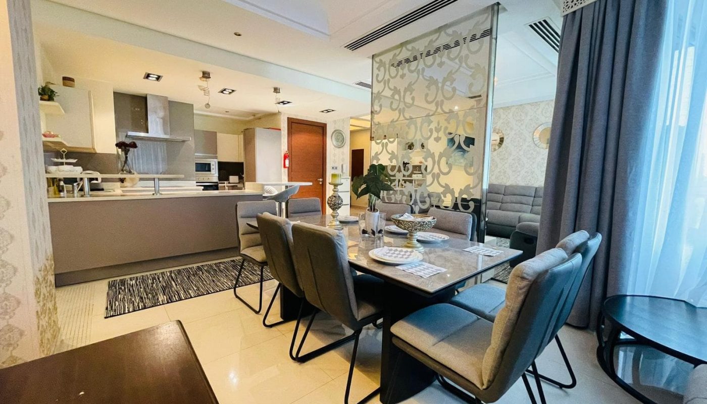 Experience the luxury of this modern open-plan dining and living area in a 2 BR apartment on Reef Island. Featuring a dining table with six chairs, a neutral-toned kitchen, and a seating area with a sofa against a decorative partition, light floods in from large windows creating an inviting ambiance.