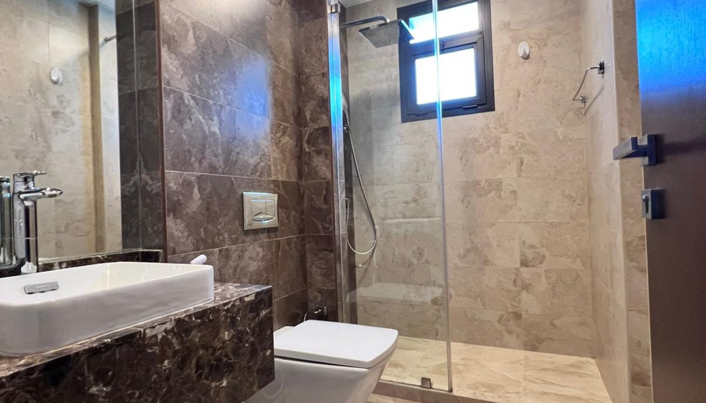 Modern bathroom with marble surfaces, a wall-mounted sink and toilet, glass-walled shower area, and a small window providing natural light.