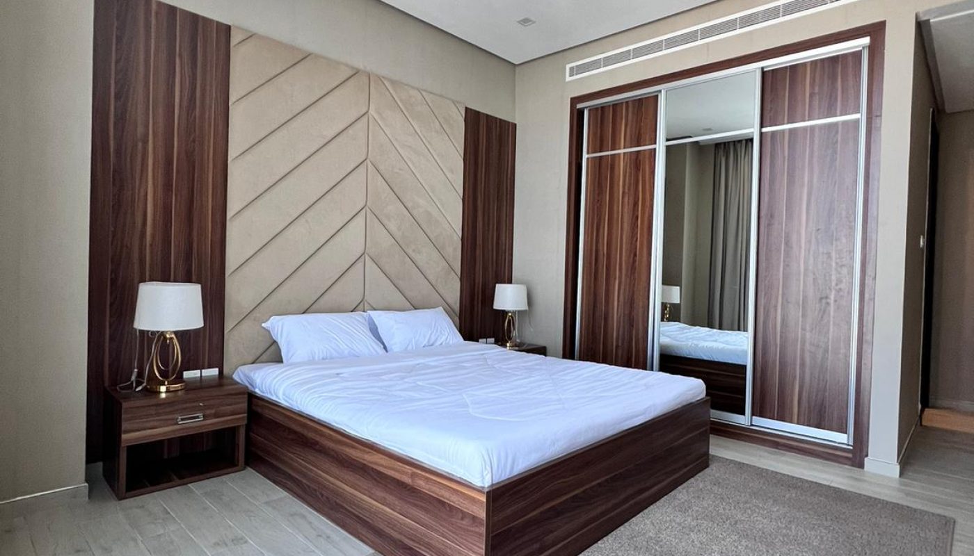 Modern bedroom with a wooden bed frame, two bedside tables with lamps, large wardrobe with sliding mirrored doors, and light-colored flooring and rug.