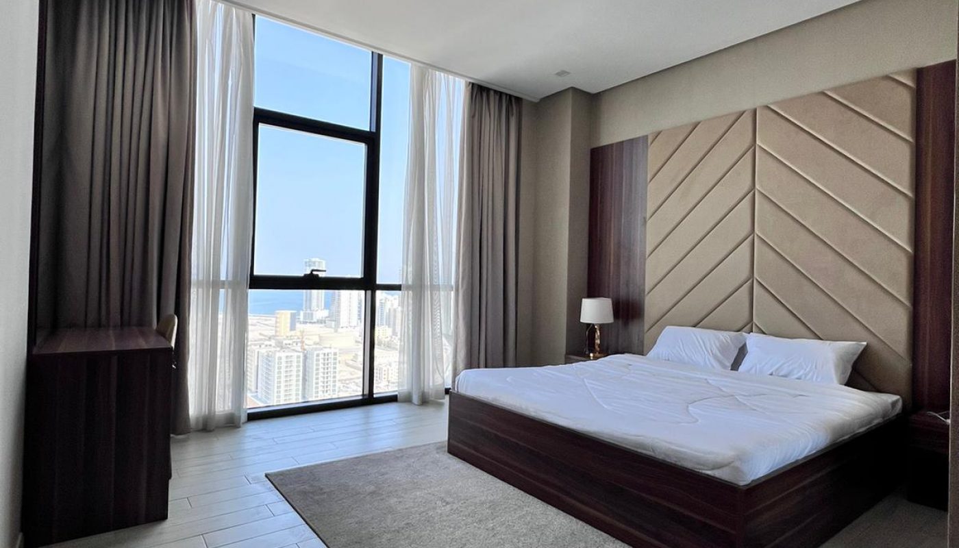 A modern bedroom with a large bed, a gray rug, floor-to-ceiling window, city view, and minimal furnishings.