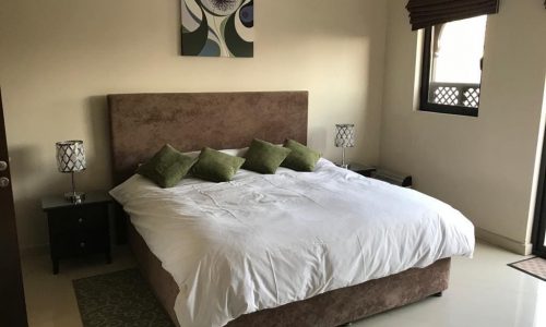 A neatly arranged bedroom featuring a large bed with a brown headboard, white bedding, green cushions, two bedside tables with lamps, a wall painting, and a window with brown blinds.