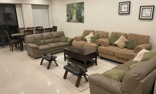 A spacious living room with two beige sofas, a brown coffee table, and a dining area in the background. The room is decorated with framed wall art and green cushions.