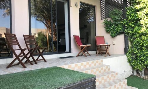 Outdoor patio with wooden chairs and a table on a tiled platform. The patio is adjacent to a building with glass doors and greenery on one side. Steps lead down to a small grassy area.