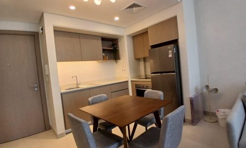 A modern kitchen with wooden cabinets, a refrigerator, an oven, and a dining table with four upholstered chairs. A ceiling light fixture hangs above the table.