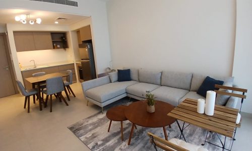 A modern, open-concept living area with a grey sectional sofa, wooden coffee tables, a dining table with chairs, and a view into a kitchen. Neutral tones and minimalist decor are featured throughout the space.