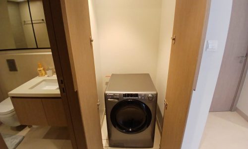 A washing machine is situated in a narrow closet with doors open. The closet is adjacent to a bathroom with a sink and mirror visible on the left.