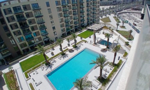 Aerial view of a modern apartment complex featuring a large rectangular swimming pool surrounded by lounge chairs, palm trees, and landscaped areas. Multiple buildings are visible in the background.