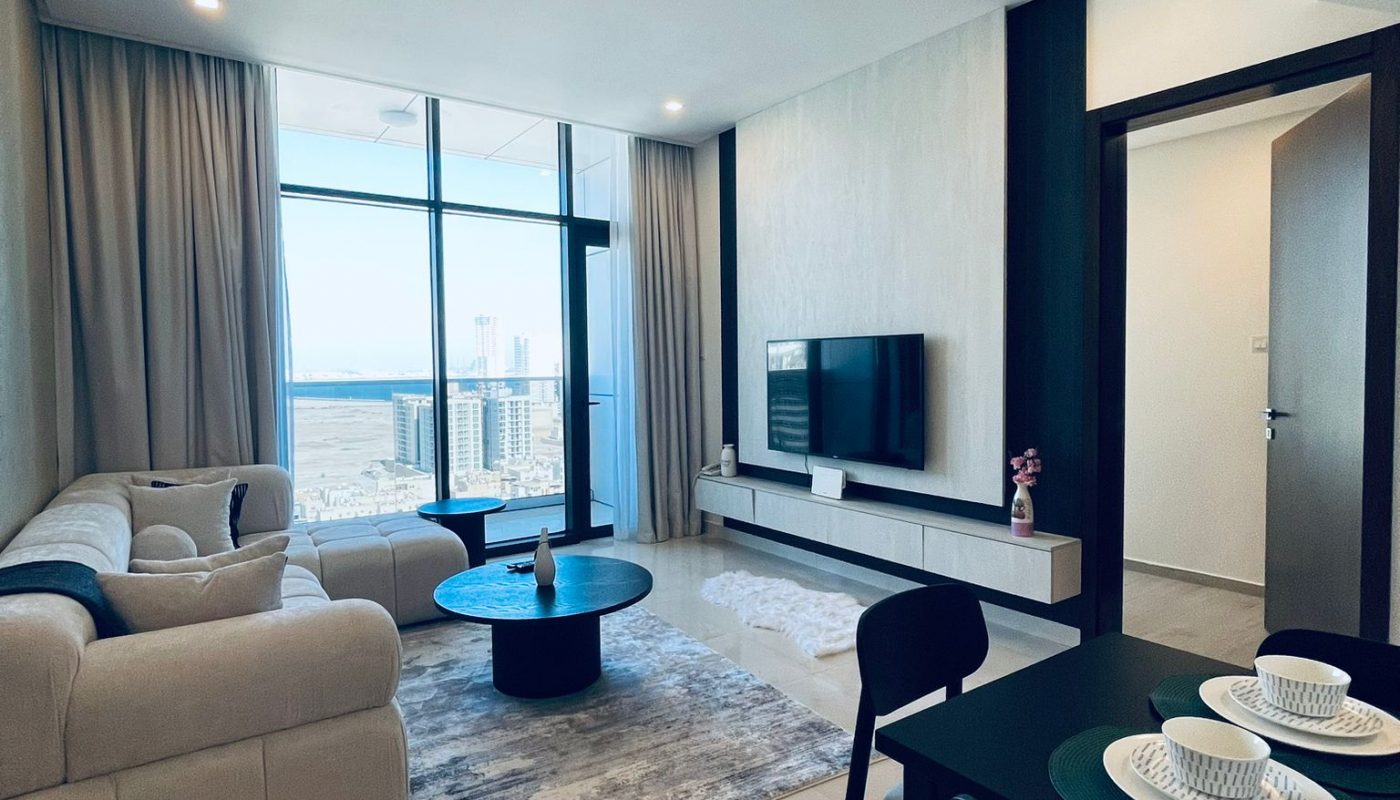 Modern living room with beige sectional sofa, rug, coffee table, and wall-mounted TV; dining table with two place settings in the foreground, large windows with city view in the background.