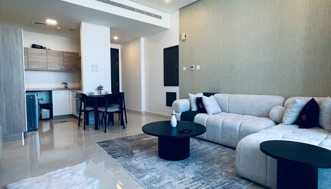 Modern living room with a light gray sectional sofa, black tables, a white faux fur rug, and an open kitchen and dining area in the background.