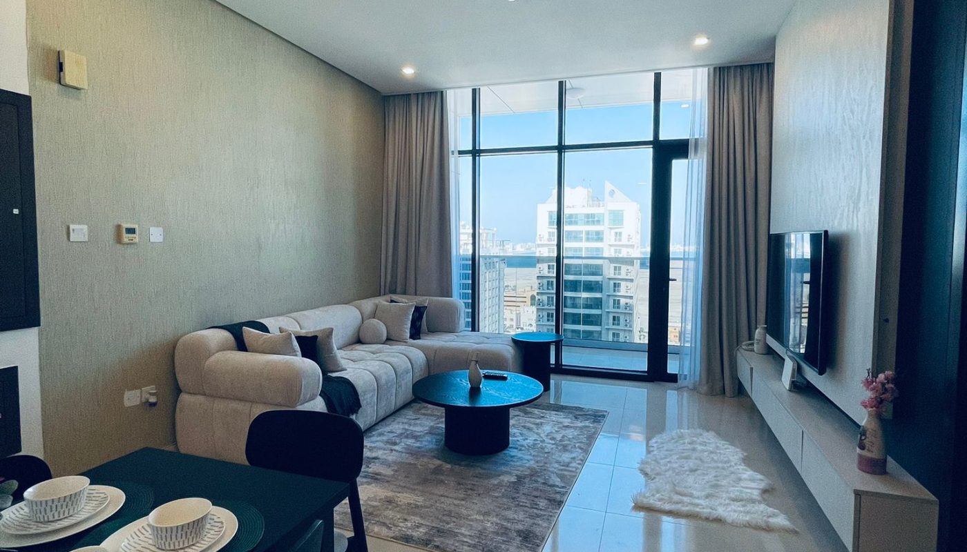 Modern living room with a beige sofa, blue coffee table, TV on a wall-mounted unit, dining table set for two, and floor-to-ceiling windows showing a cityscape view.
