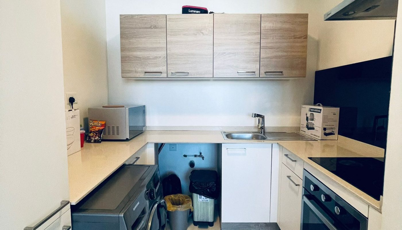 Modern compact kitchen with white cabinets, wooden upper cupboards, washing machine, oven, sink, microwave, and small appliances on the countertop. Trash bins are under the counter.
