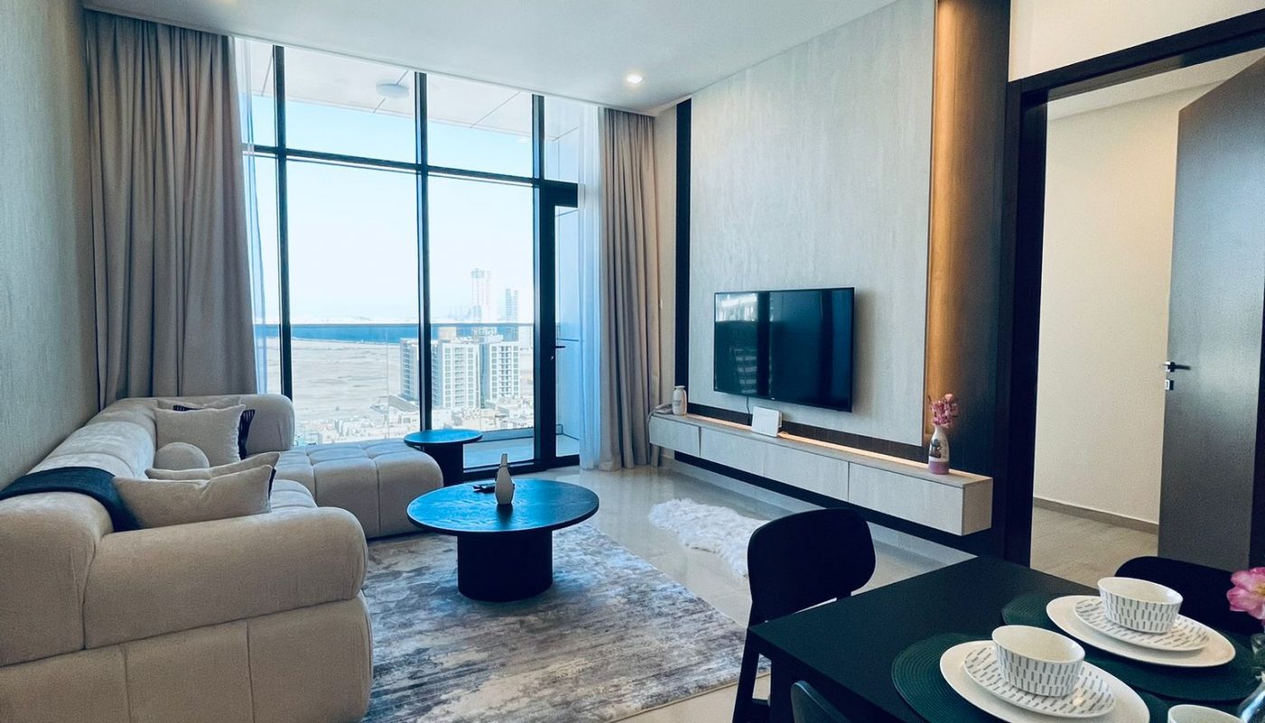 Modern living room with a beige sofa, coffee table, TV on the wall, dining table set for three, and large windows overlooking a cityscape.