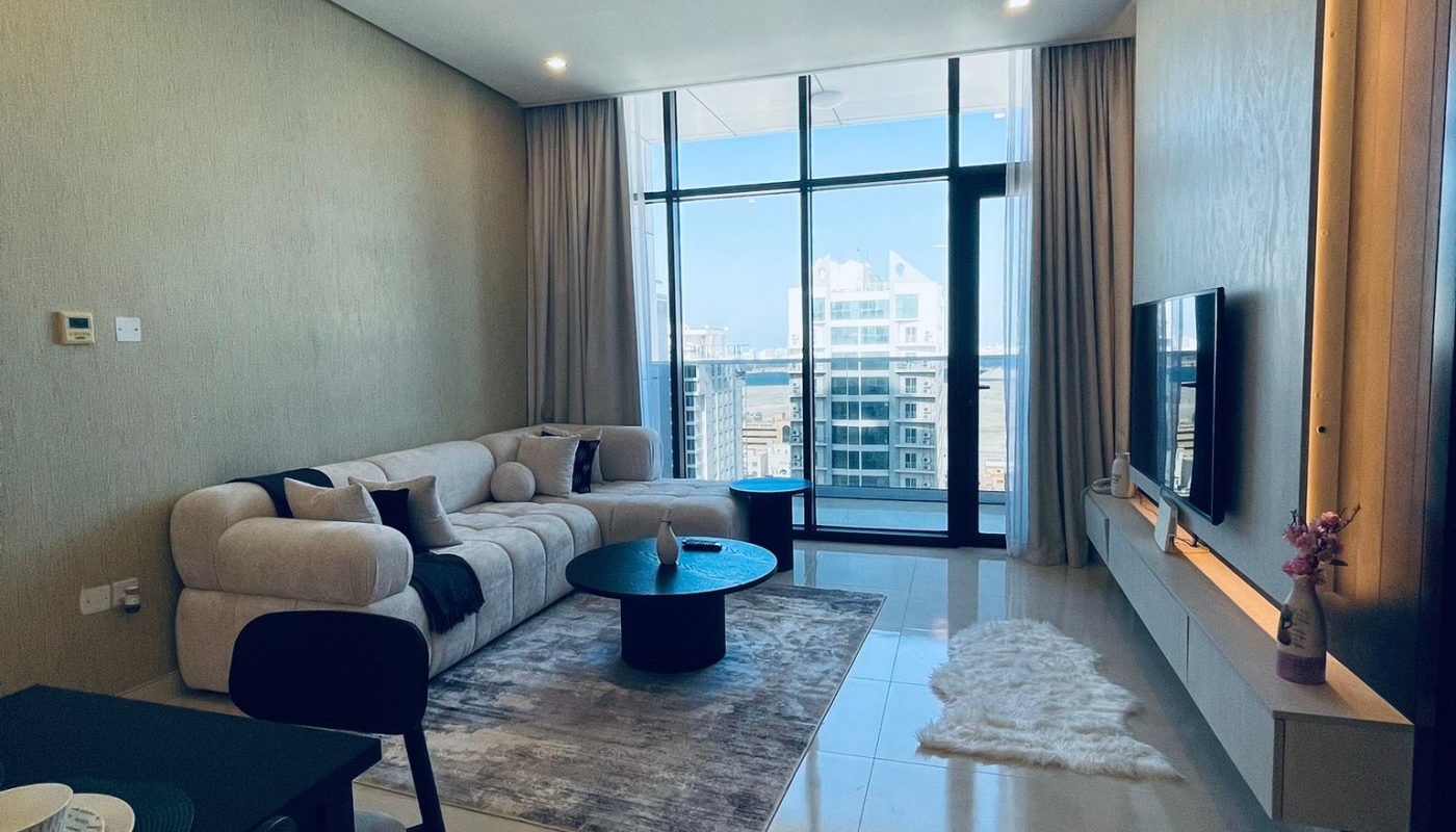 Modern living room with a beige sofa, coffee table, wall-mounted TV, large windows with city view, and neutral decor. Dining area is partially visible in the foreground.